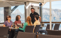 Vier Personen sitzen an einem Tisch auf einer Sonnenterasse mit Bergblick und werden von einem lächelnden Kellner bedient | © Bergbahnen Flumserberg AG