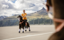 Frau sitzt auf einem Spielzeugpferd mit Rollen und fährt auf einer Fläche, umgeben von einer Berglandschaft | © Bergbahnen Flumserberg AG