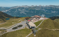 Luftaufnahme einer Bergstation mit Restaurant und Seilbahn, umgeben von grünen Hügeln und einem Bergpanorama | © Bergbahnen Flumserberg AG