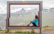 Frau sitzt in einem grossen Holzrahmen und blickt auf eine Berglandschaft | © Bergbahnen Flumserberg AG