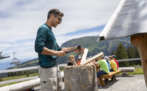 Mann hackt ein Holzstück mit einer Axt an einer Feuerstelle | © Bergbahnen Flumserberg AG