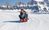 Erwachsene Frau und Kleinkind auf einem Schlitten, gleiten eine verschneite Piste hinunter | © Bergbahnen Flumserberg AG