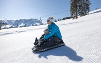 Erwachsene Person mit Kind auf dem Schlitten, rutschen bei sonnigem Wetter eine verschneite Piste hinunter | © Bergbahnen Flumserberg AG