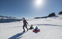 Erwachsene Person zieht zwei Kinder auf Schlitten über eine verschneite Piste bei sonnigem Wetter | © Bergbahnen Flumserberg AG